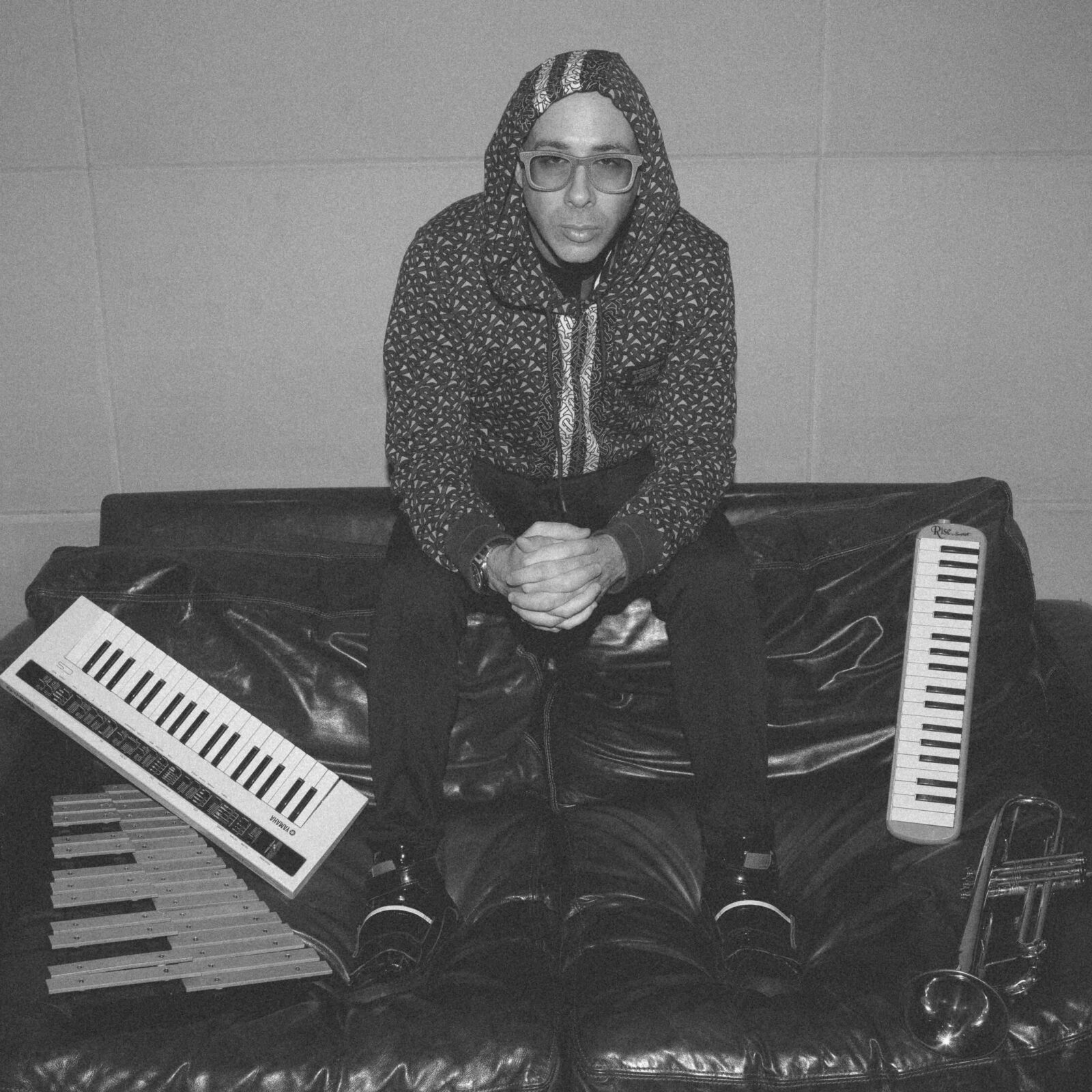 Black and white portrait of musician Sam Barsh sitting on top of a coach with two keyboards and a trumpet at his side.
