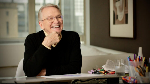 A portrait of American fashion designer and costumier sitting at his desk, smiling.