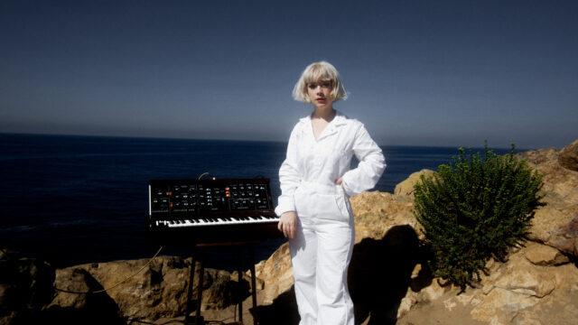 Danz CM wearing a white boiler suit, standing in front of a synth.