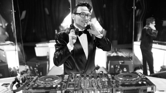 Black-and-white portrait of DJ Johnny Dynell standing behind a DJ booth, dressed in a tuxedo and bow tie.