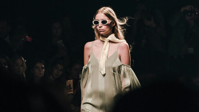 A runway model walks toward the camera wearing a pale green, flowing dress with thin straps and draped sleeves, styled with a cream scarf tied at the neck and oval white sunglasses, against a darkened audience backdrop.