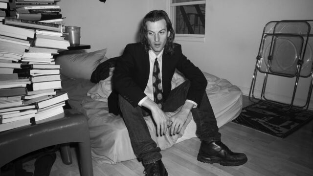 A black and white portrait of the actor, writer and filmmaker Peter Vack sitting on his bed with a pile of books nearby.
