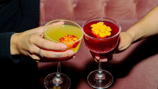 Two people clink colorful cocktails in coupe glasses, each topped with an edible flower.
