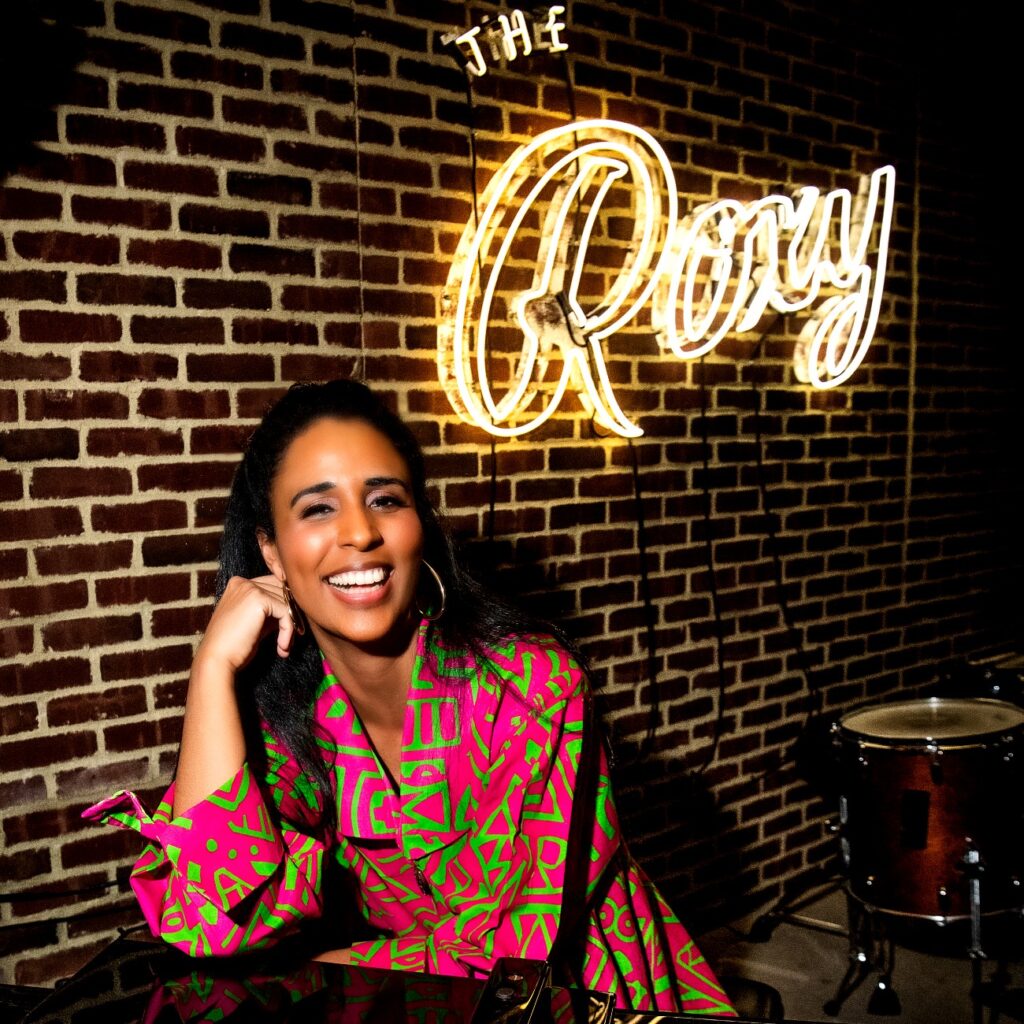 Singer Camila Cortina sitting with one elbow on a piano at The Roxy Hotel.