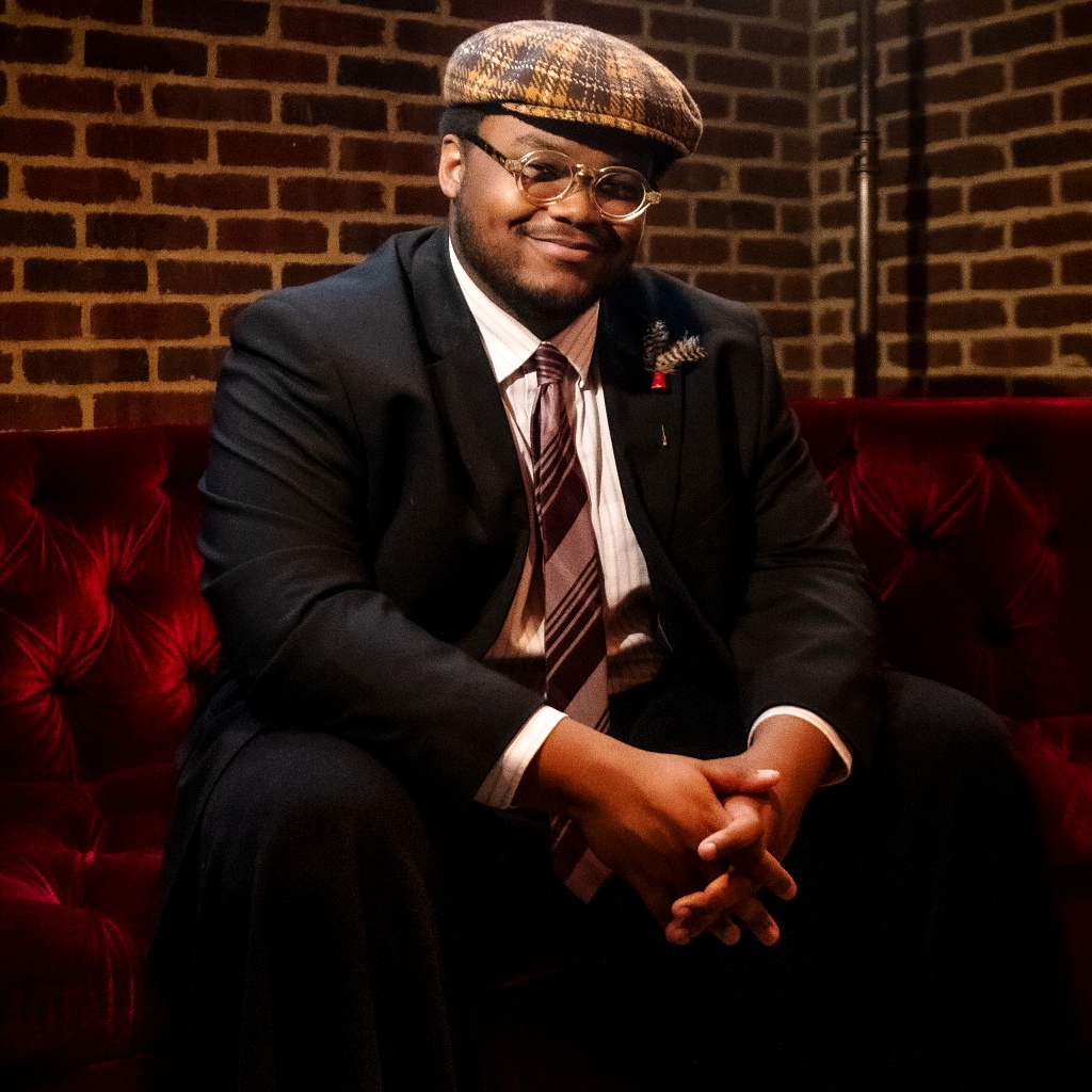 Musician William Hill III sitting on a red velvet couch wearing a suit.