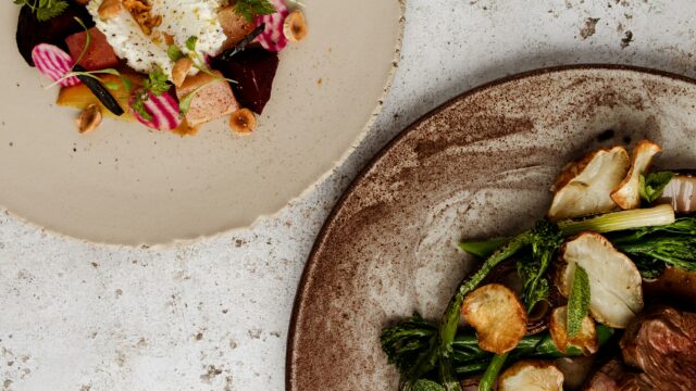 Overhead of two plated dishes: roasted vegetables with sliced meat on a brown plate, and a colorful beet, radish, and cheese arrangement on a light plate.