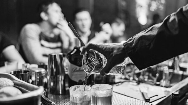 Bartender pouring cocktails into two short glasses across a busy New York bar counter, with patrons blurred in the background.