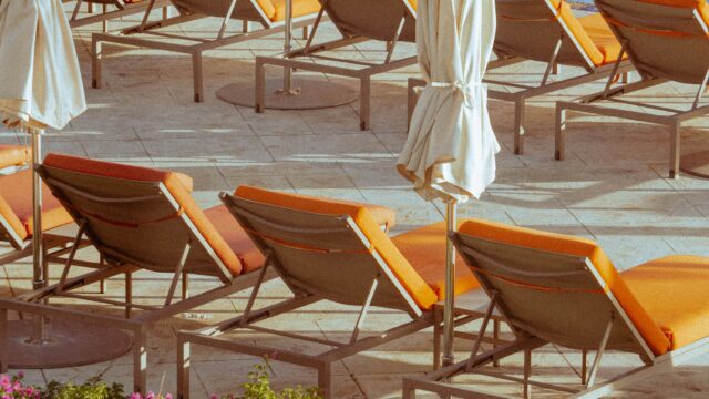 Rows of deck chairs with with orange cushions arranged on a stone pool deck, interspersed with closed white canvas umbrellas in warm golden sunlight, with pink flowering shrubs visible in the foreground.