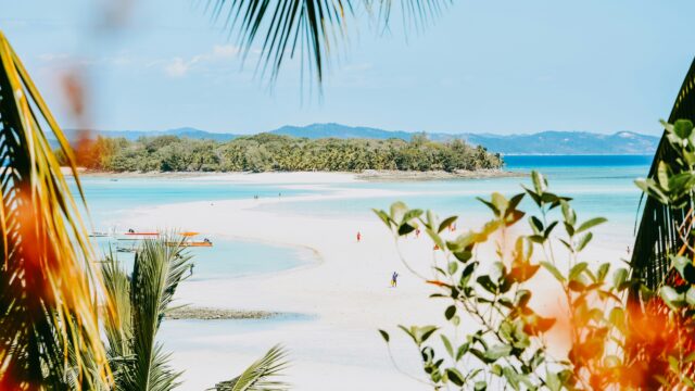 A tropical white sand beach with turquoise water, framed by palm leaves.