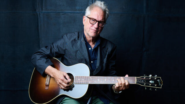 Gerry Beckley, co-founder of America, holding an acoustic guitar and looking at the camera.