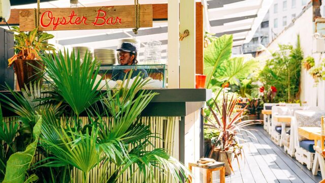 A bartender at the Gilligan's Oyster Bar, which is decorated with white painted bamboo, palm leaves and a wooden sign that reads "Oyster Bar" in red, cursive writing