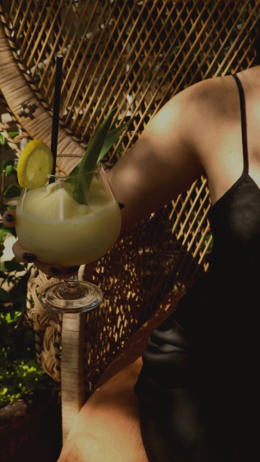 Woman sitting on a wicker chair holding a frozen cocktail and wearing a black slip dress.