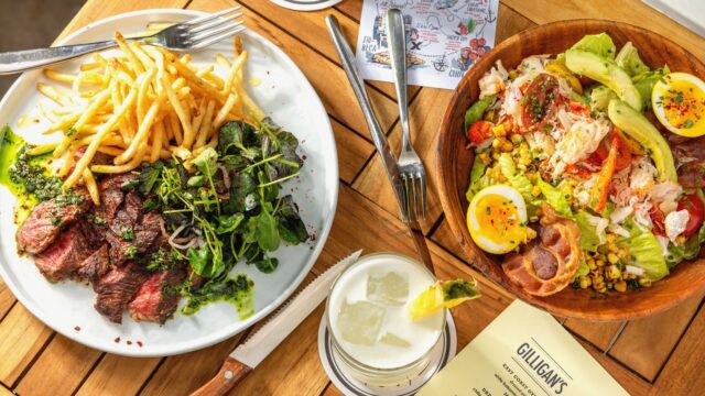 Steak entree with fries and a Cobb Salad on a wood table at Gilligan's outdoor restaurant in SoHo, NY.