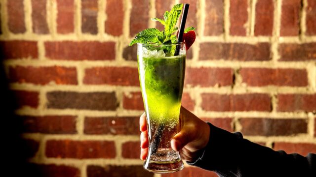 Man holding up a green matcha iced beverage with green garnish and black straw in front of a brick wall.