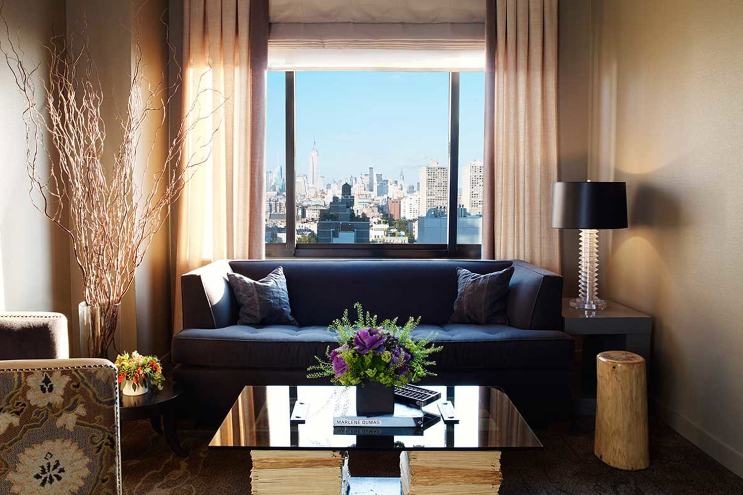 Deluxe One Bedroom Suite living rooo with a blue couch, coffee table with flowers and a plush chair. there is a view of the manhattan skyline