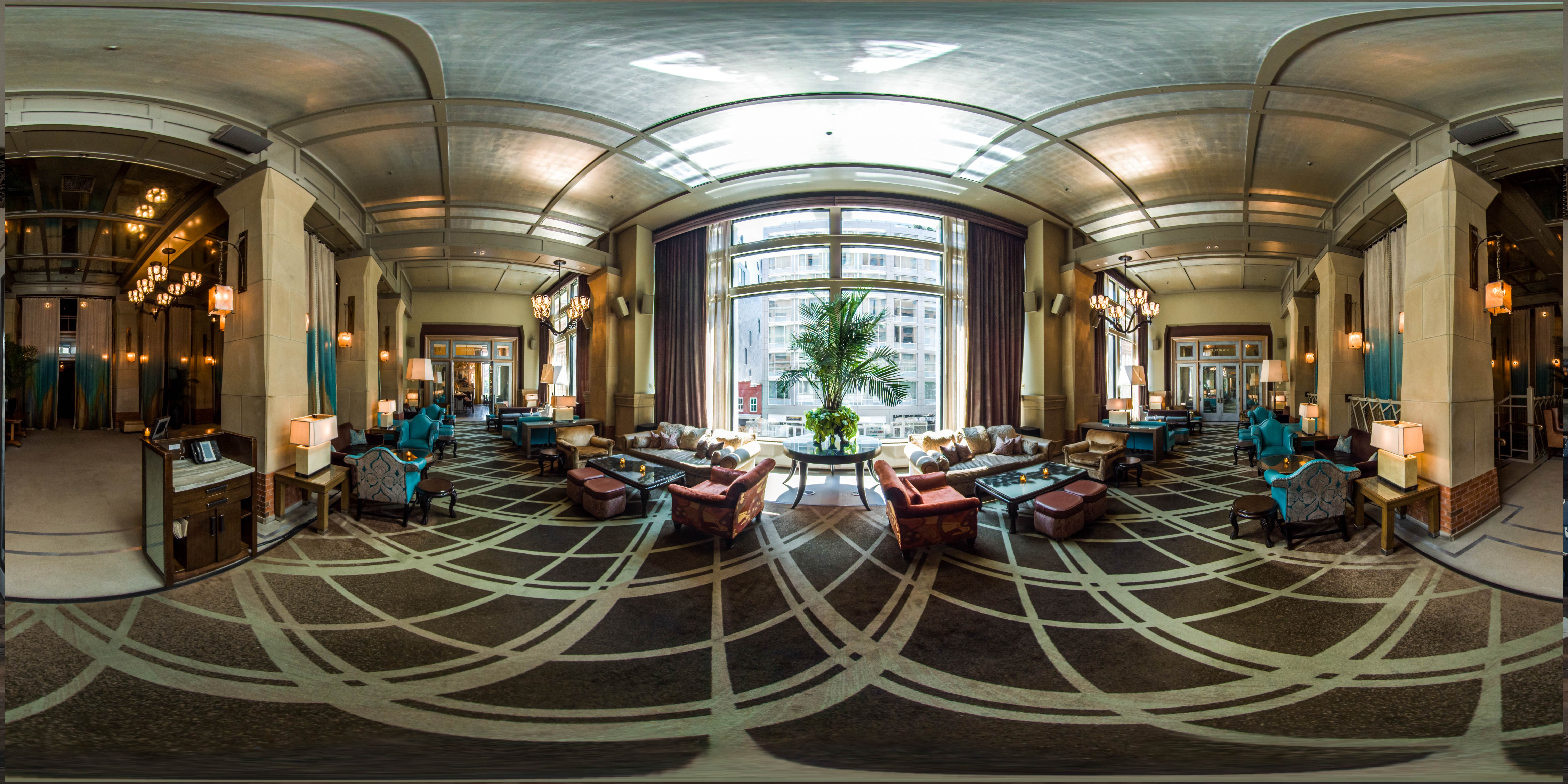 A wide-angle view of a luxurious hotel lobby with a high ceiling, large window, seating areas, and decorative lighting.