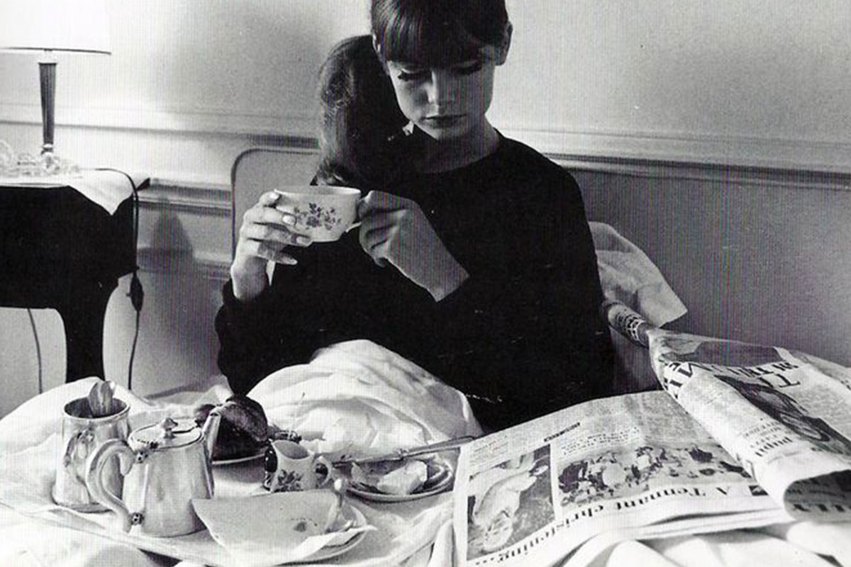 black and white image of a woman laying in bed with breakfast and a newspaper
