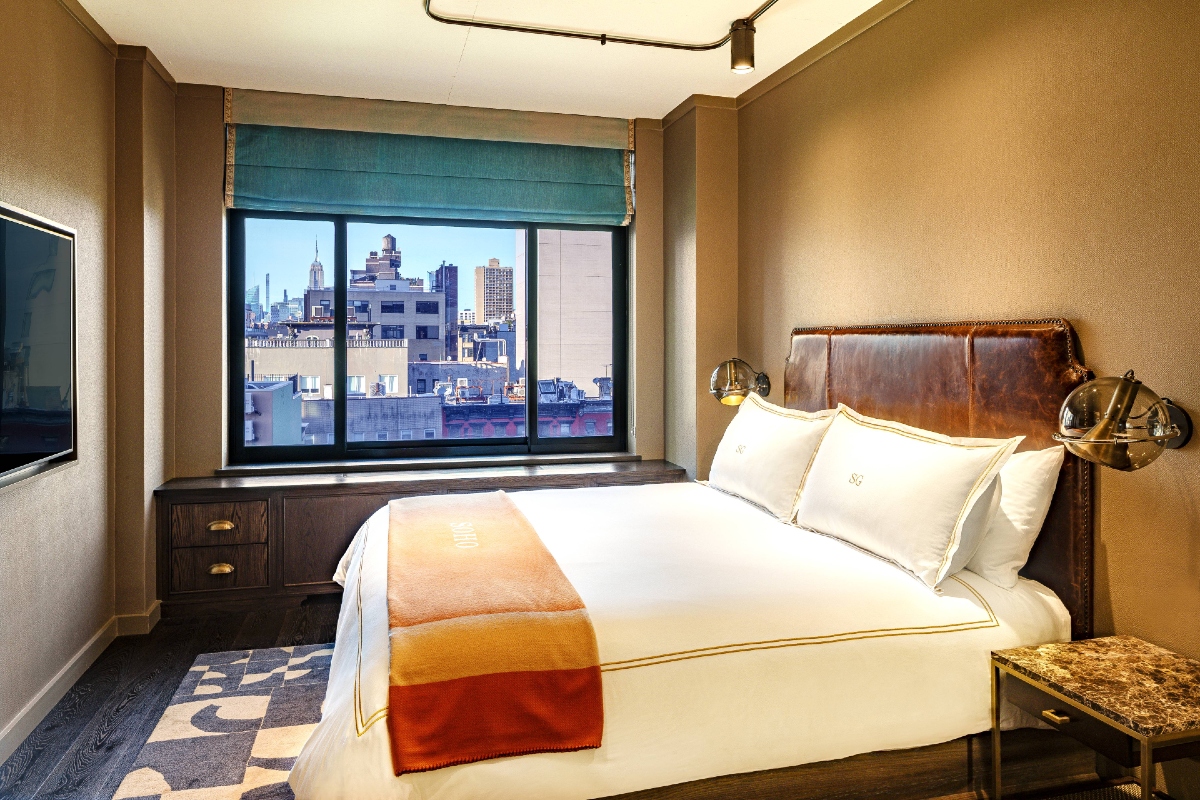King bed with white sheets, orange throw blanket, and brown headboard with window looking north over the Manhattan skyline from Soho Grand Hotel.