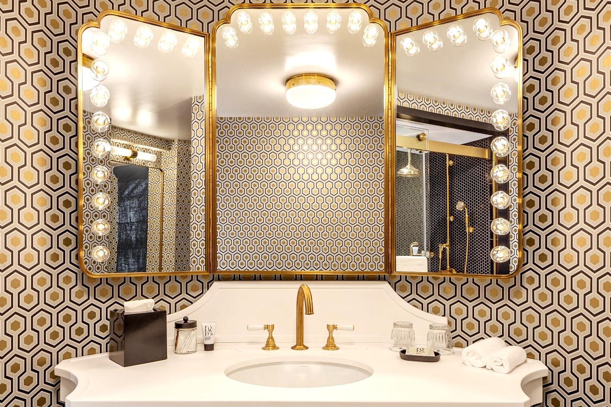Vanity closeup of triptych mirror, surrounding bulb lighting, white sink and brass plumbing finishes.