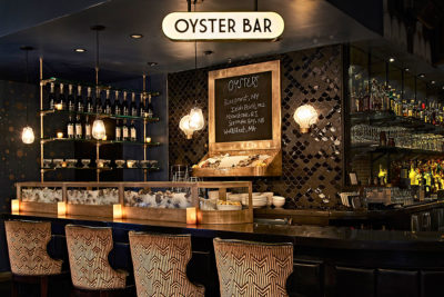the oyster bar. there are shelves of liquor bottles, a menu board and oysters over ice on display