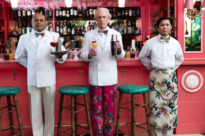 three men in kitchen whites stand in front of a red bar holding different craft cocktails