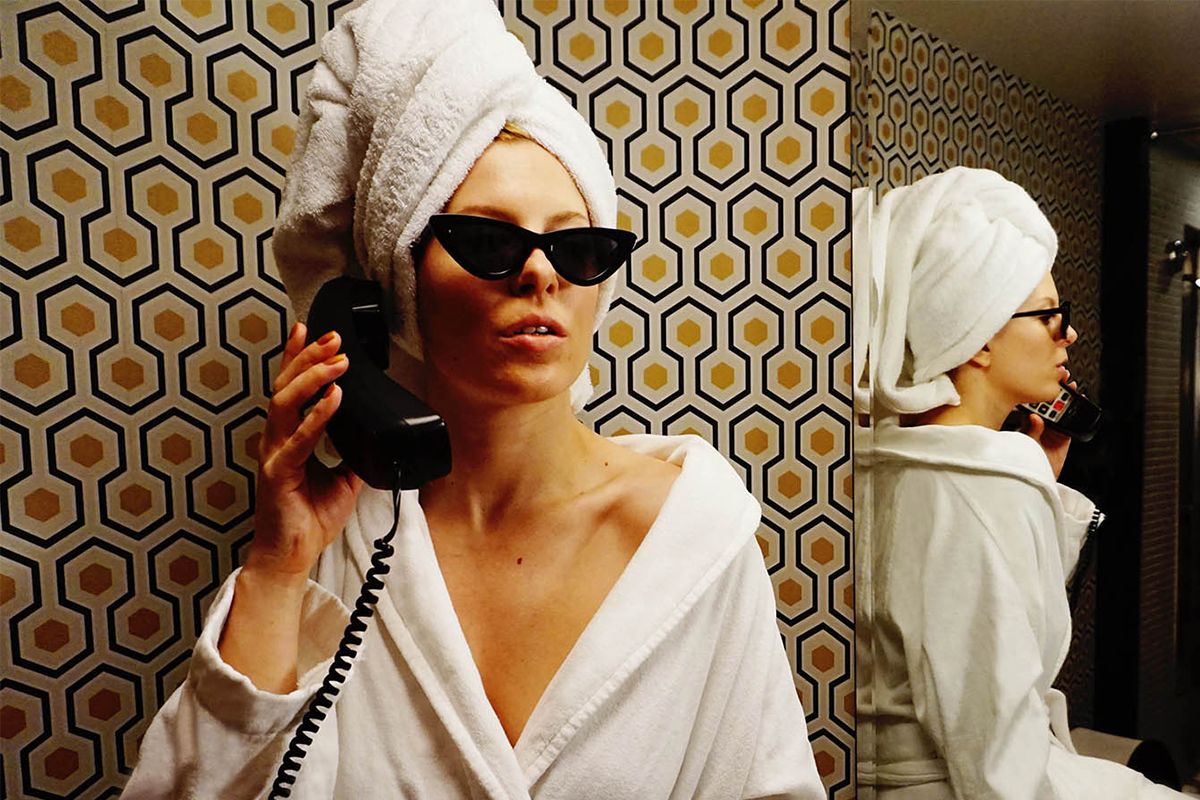 a woman in a robe and sunglasses stands in a Roxy Hotel bathroom talking on a retro telephone