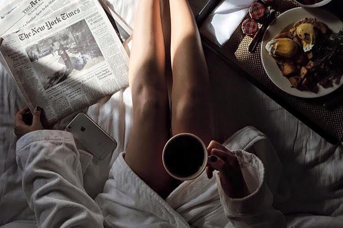 a woman sits on a Roxy Hotel bed in a robe enjoying room service and reading the complimentary New York Times