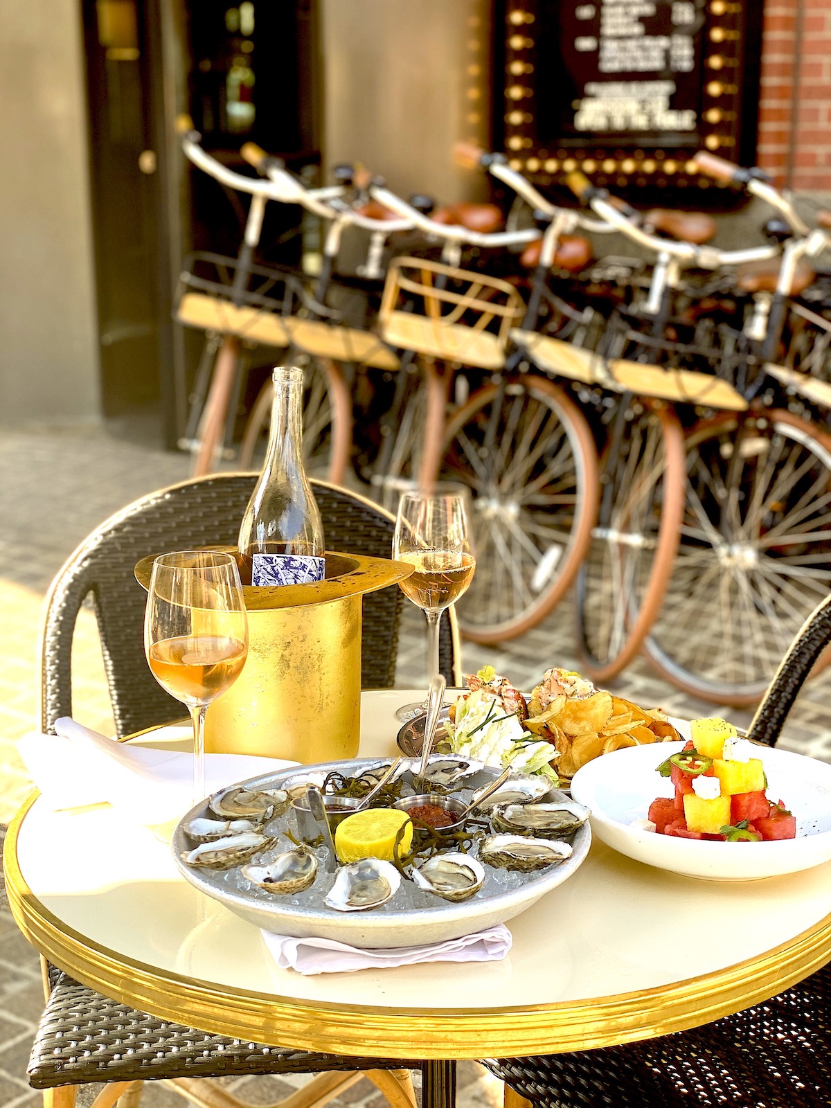 a spread of oysters, fruit and chips at an outdoor table. there is champagne chilling with two glasses next to it