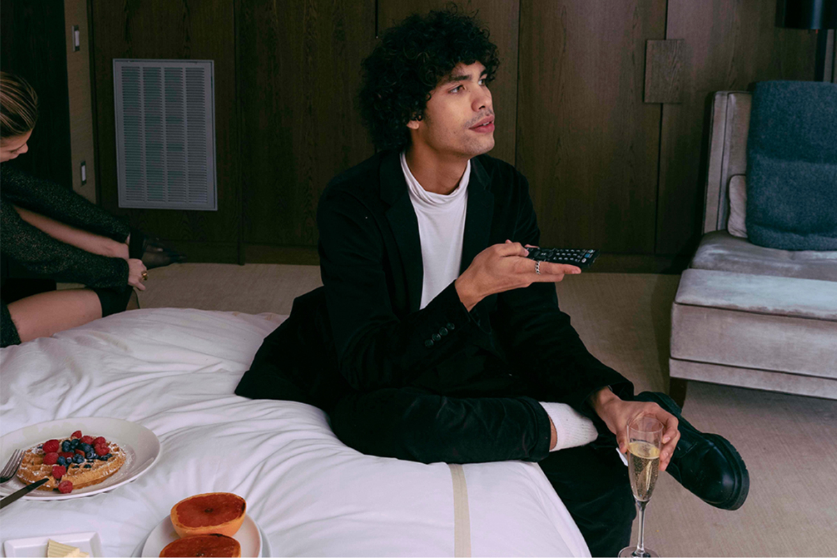 a well dressed man sits on the edge of a Roxy Hotel bed using a remote to watch television