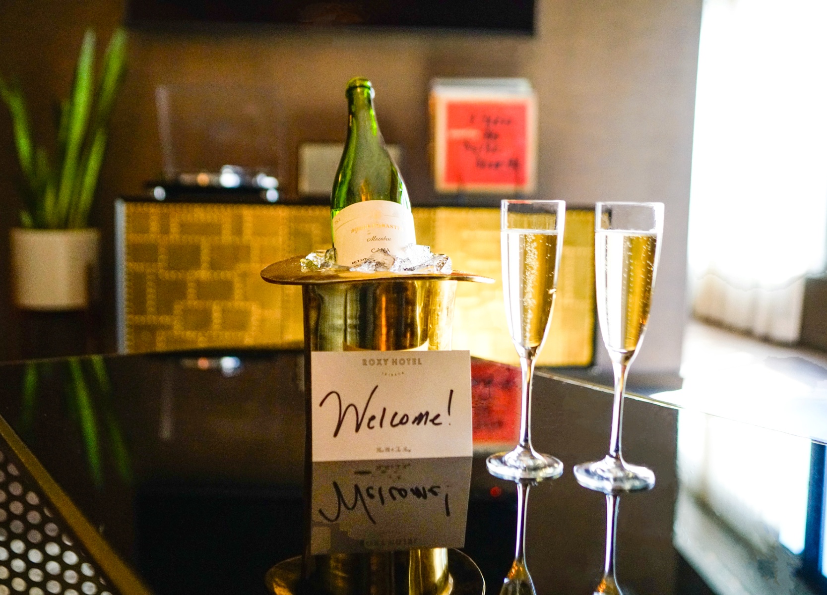 Two champagne flutes on a table next to a champagne bottle on ice an a welcome sign.
