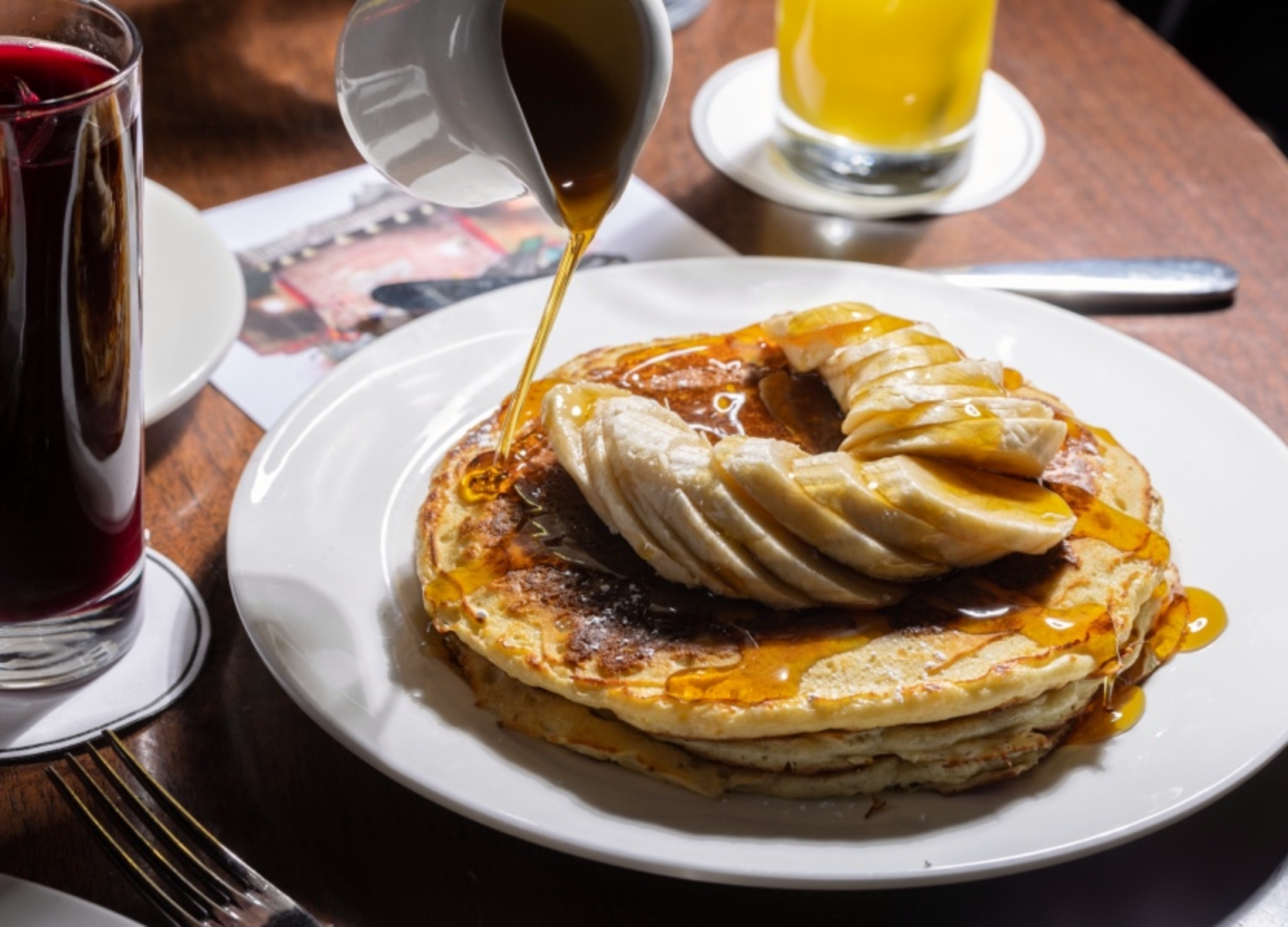 Maple syrup being poured over pancakes with slices of bananas on top.
