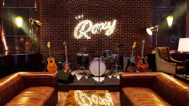 The Roxy Bar stage. a brick wall sits behind instruments. a neon roxy sign hangs abut the stage