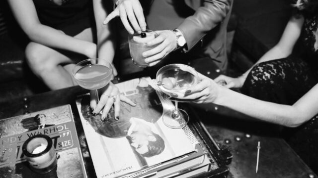 black and white image of three friends dressed in formal attire raising their craft cocktails in a cheers