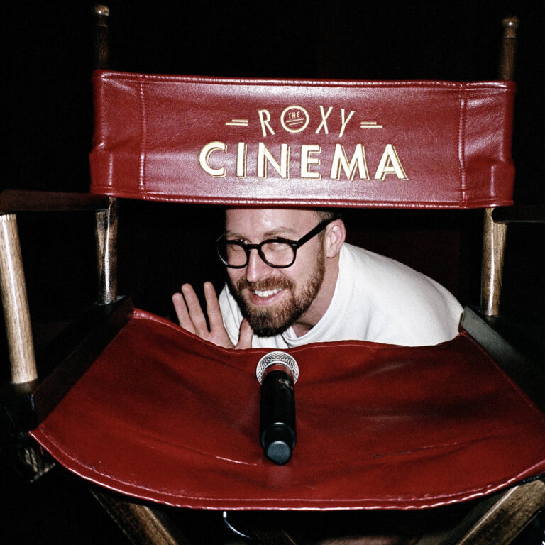 a man crouches behind a red leather roxy cinema new york directors chair and smiles and waves