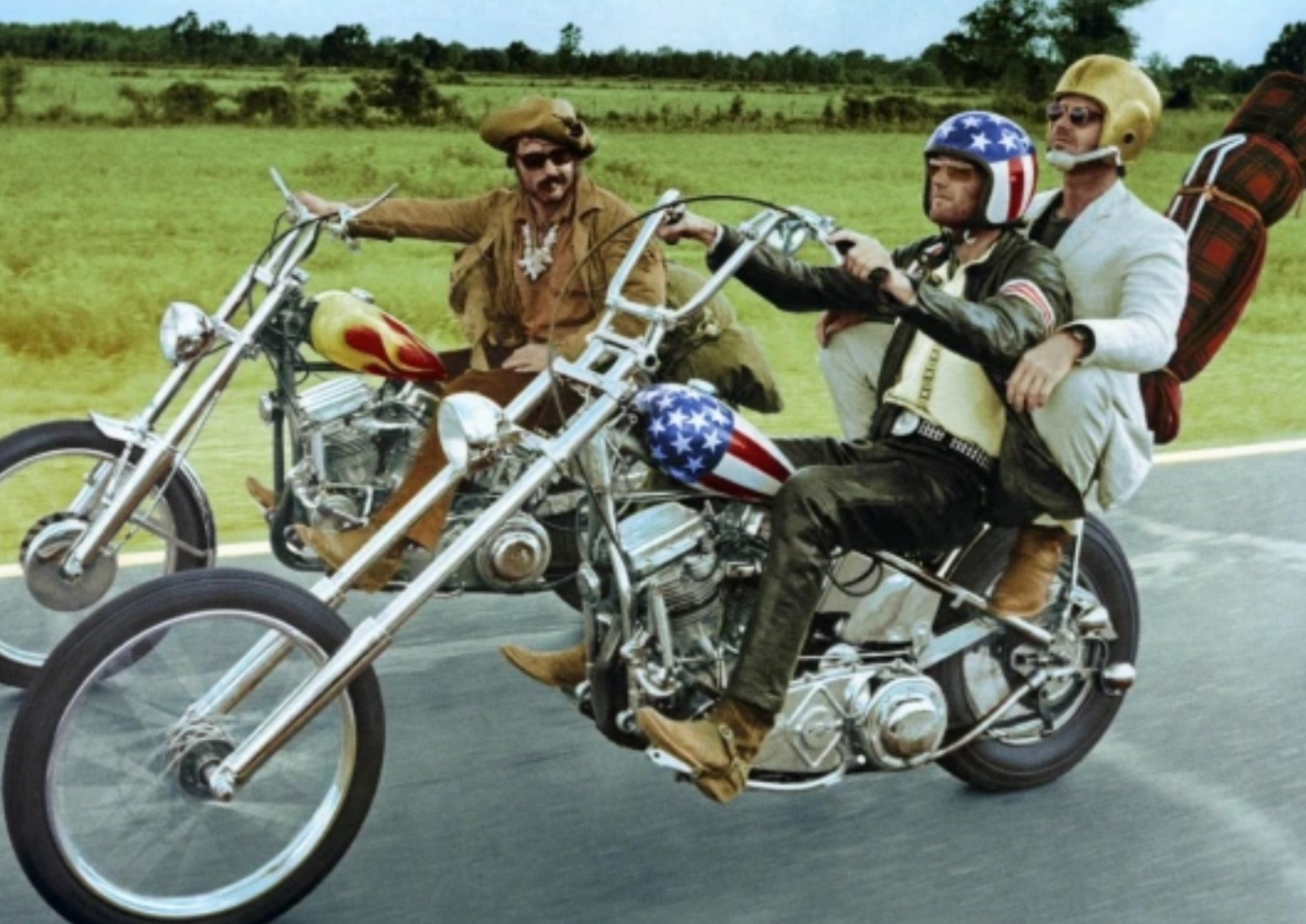 three men riding on motorcycles. one wears an american flag patterned helmet to match his bike and another is dressed as a cowboy