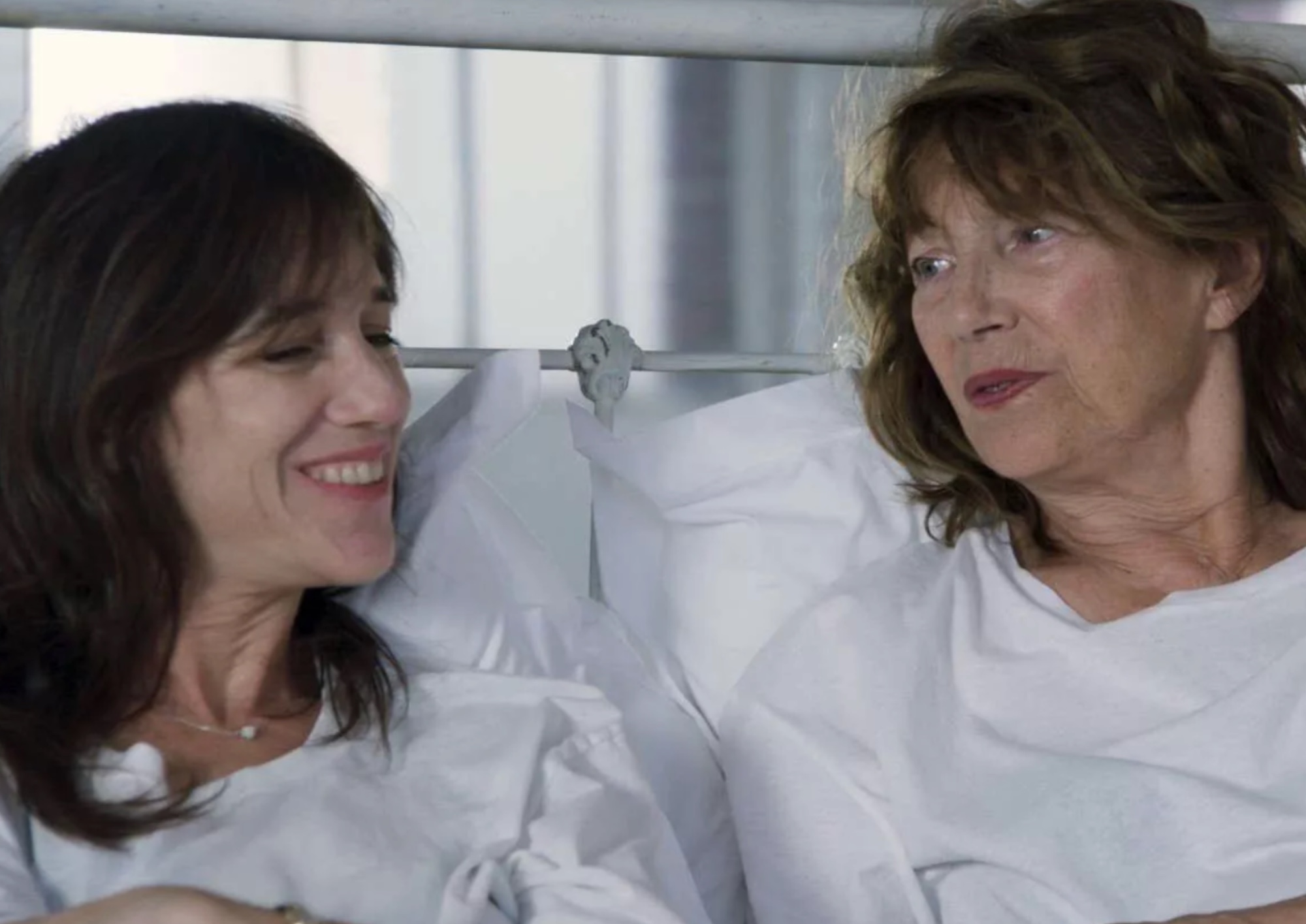 an image of two women in white sit on a bed and talk to each other