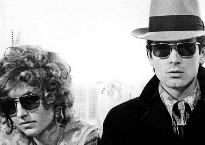 a black and white photograph of a man and a woman wearing disguises walking towards the camera . the skyline of a city is in the background