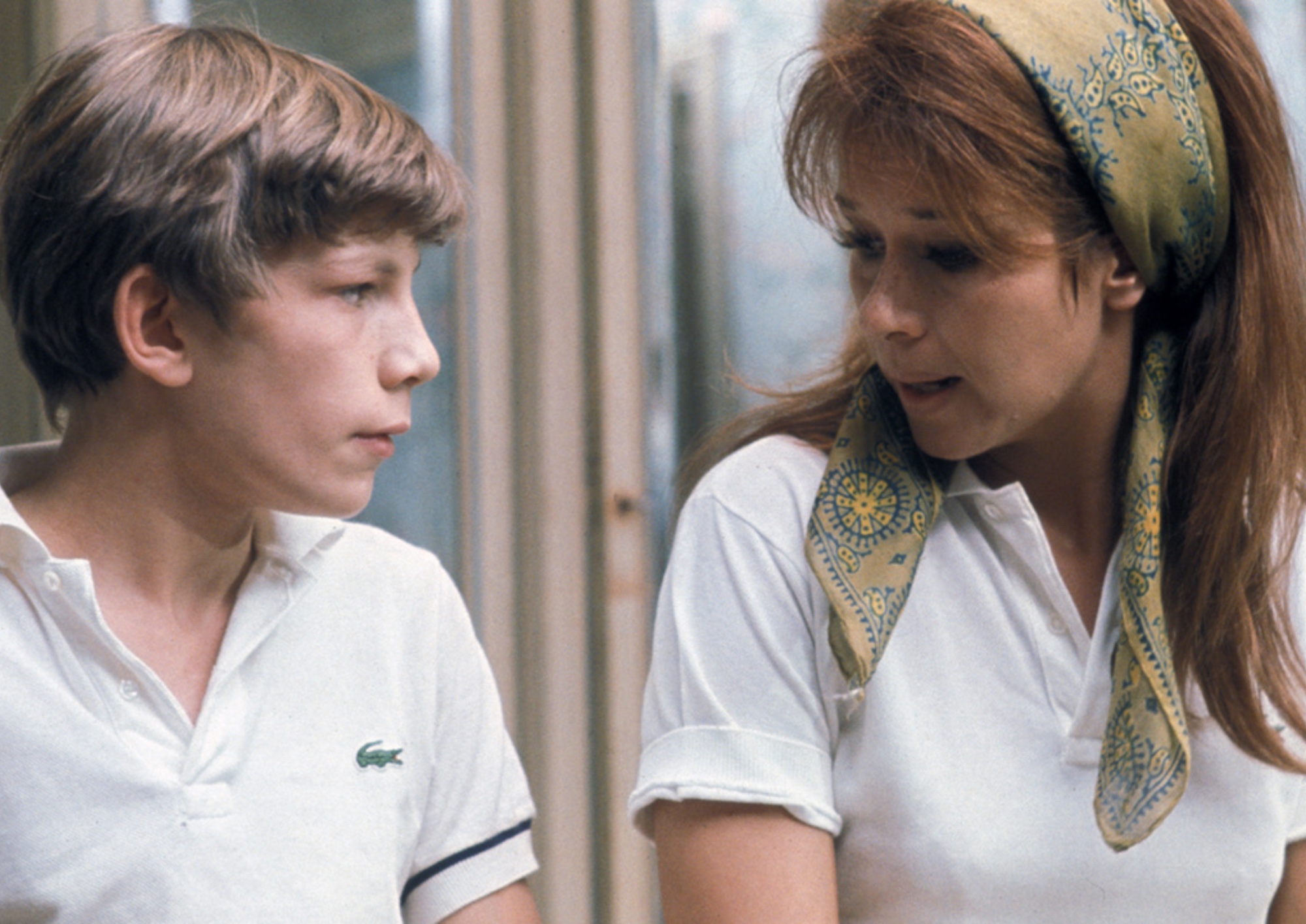 a woman with a white shirt and colorful hair scarf talks to a young boy with a white collared shirt