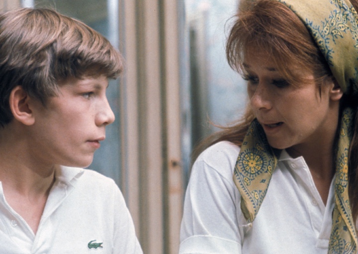 a woman with a white shirt and colorful hair scarf talks to a young boy with a white collared shirt