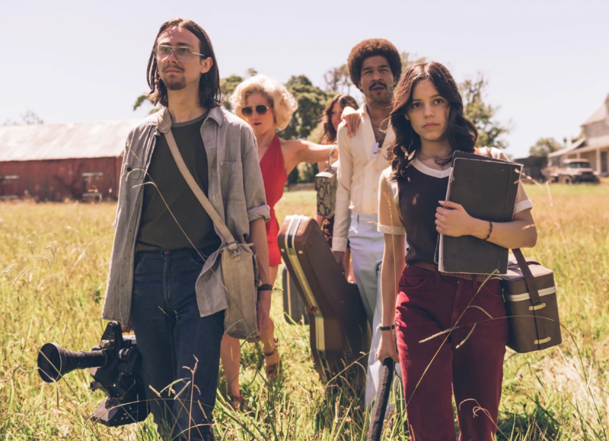 five people dressed in 70s attire and carrying film-making gear are walking in a rural field with tall grass.