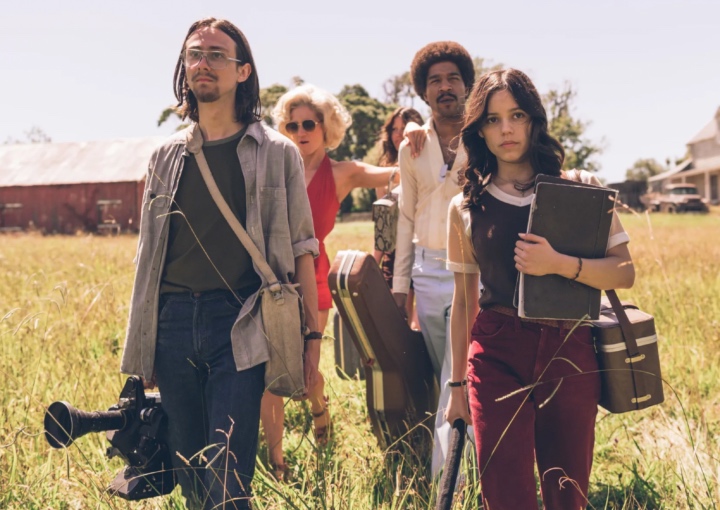 five people dressed in 70s attire and carrying film-making gear are walking in a rural field with tall grass.