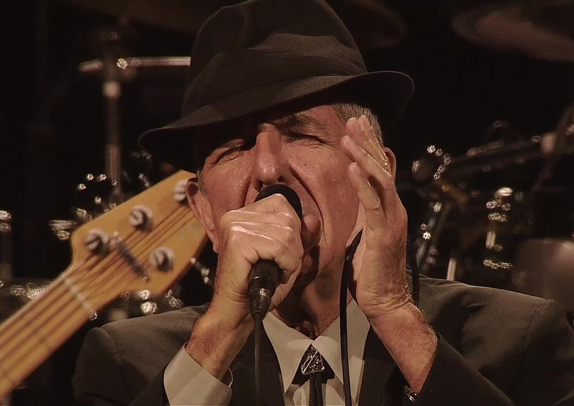 Leonard Cohen singing into a microphone with eyes shut and wearing a hat.